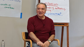 Photo shows Robert Gonzales sitting in a chair, leading a workshop. He is dressed casually in a burgundy shirt and has a microphone pinned to his shirt. Behind him are two flip charts with handwritten notes.